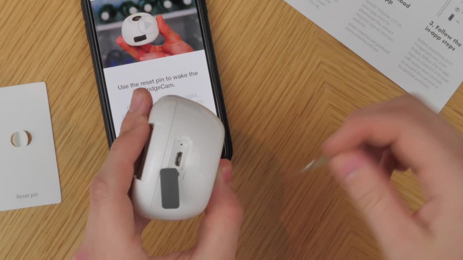 FridgeCam setup instructions on a mobile phone alongside the physical device and reset pin.